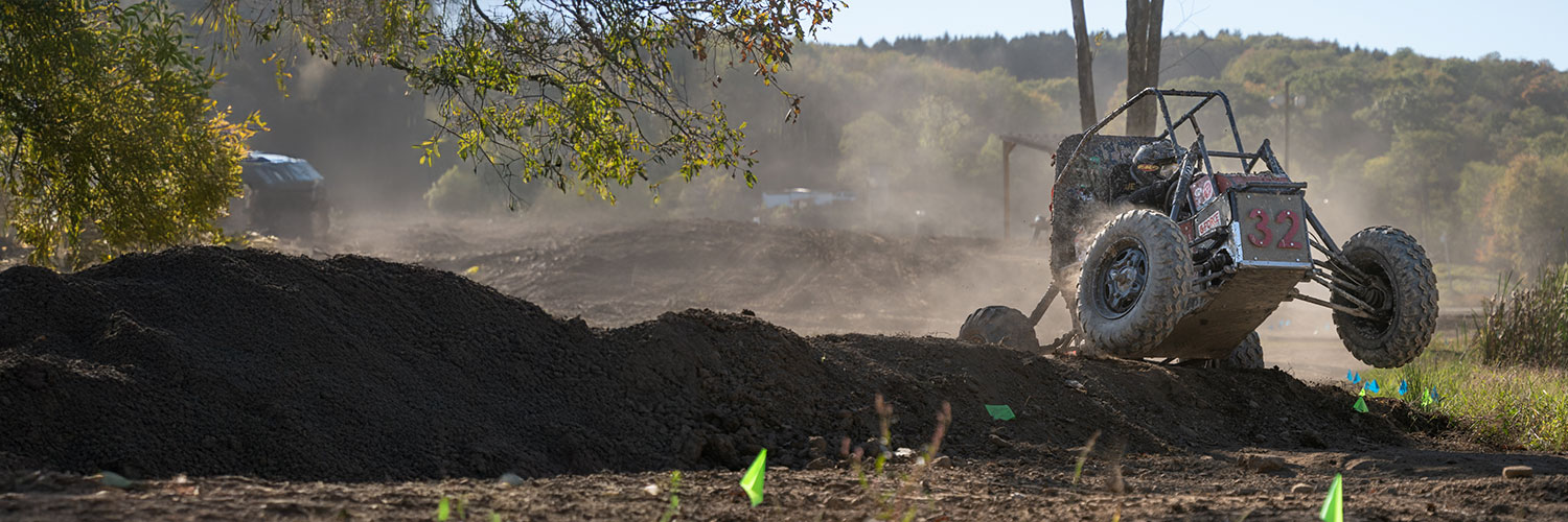 A construction site with a dirt mound in the foreground and a tree branch overhead. Dust rises in the background where machinery is at work. A small green flag is visible on the ground, and a forested hillside is in the distance.
